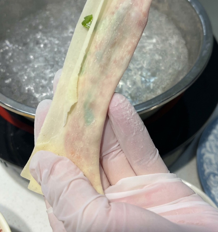 끓는 물이 담긴 냄비 위에서 장갑을 낀 두 손으로 속을 채운 완탕피를 길고 얇고 투명한 면 모양으로 늘리는 모습.