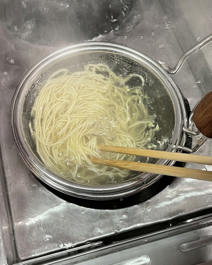 둥근 매쉬 면 건지기 채망에 담긴 얇고 연한 밀가루 면발을 나무 젓가락으로 저으며 끓는 물에서 건져 올리는 모습.