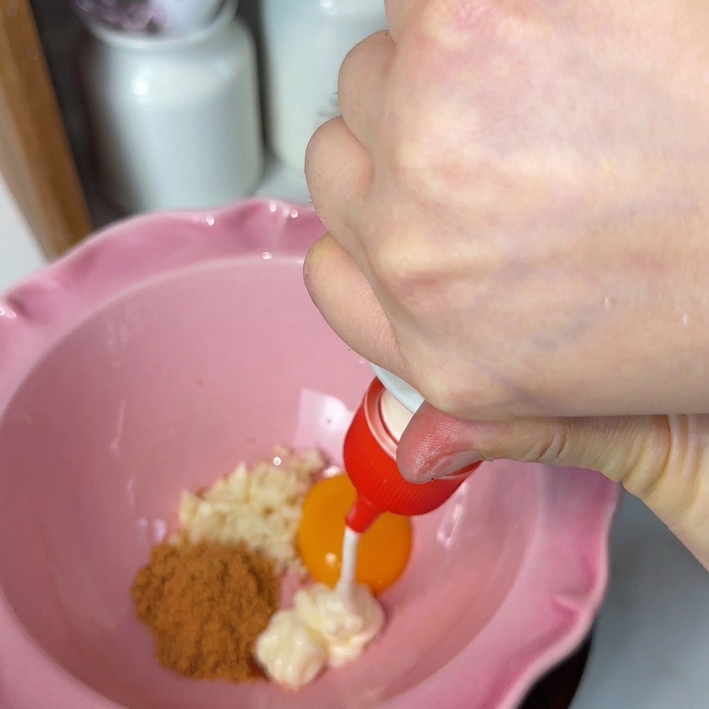 다진 마늘, 주황빛 계란 노른자, 양념 가루가 담긴 분홍색 그릇에 마요네즈를 짜넣는 모습.