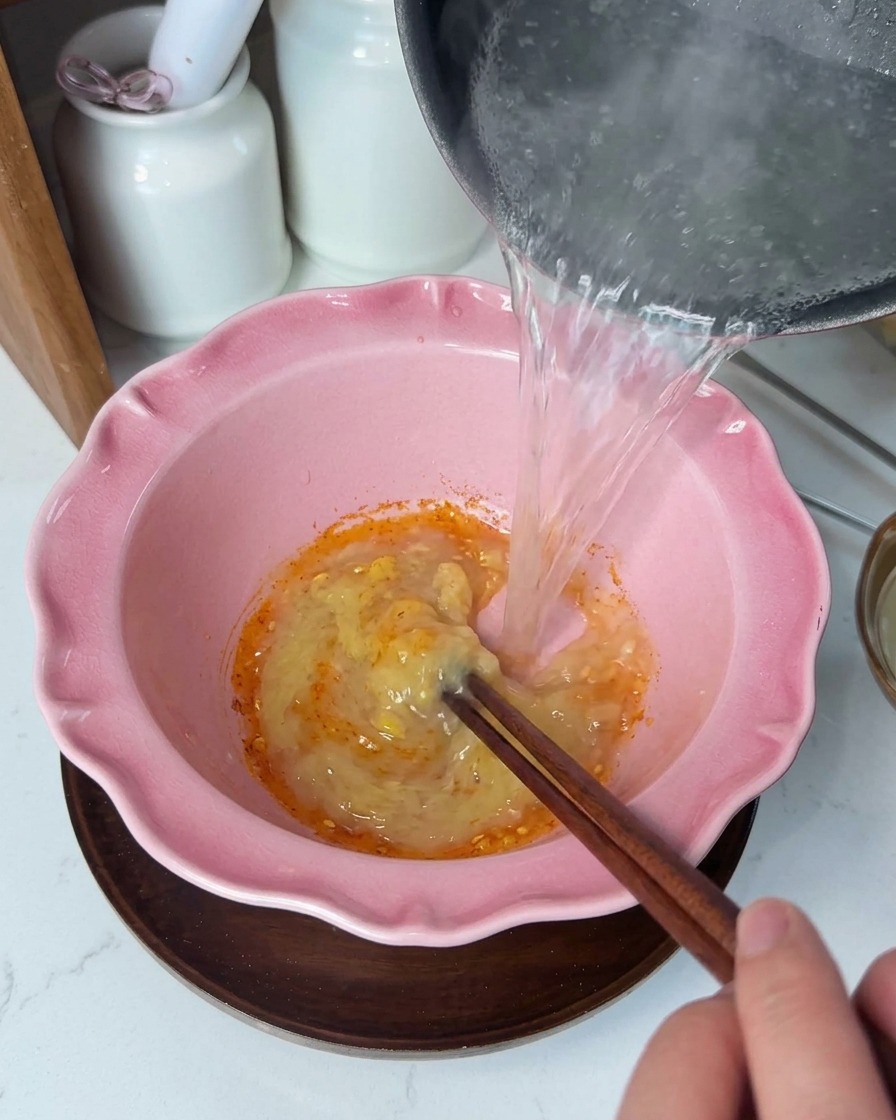 노란색 양념 페이스트가 담긴 분홍색 그릇에 뜨거운 맑은 육수를 붓고 젓가락으로 젓는 모습.