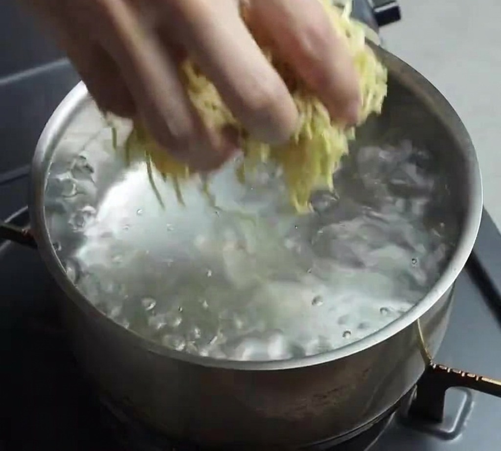 팔팔 끓는 물이 담긴 스테인리스 냄비에 생라멘 노란 면발 한 뭉치를 넣는 손.