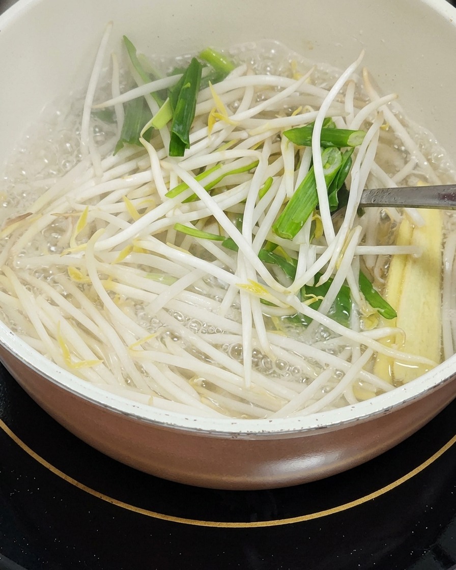 숙주나물, 썰어둔 풋마늘 잎, 영콘 조각이 담긴 냄비 안을 금속 조리도구로 젓고 있는 모습.