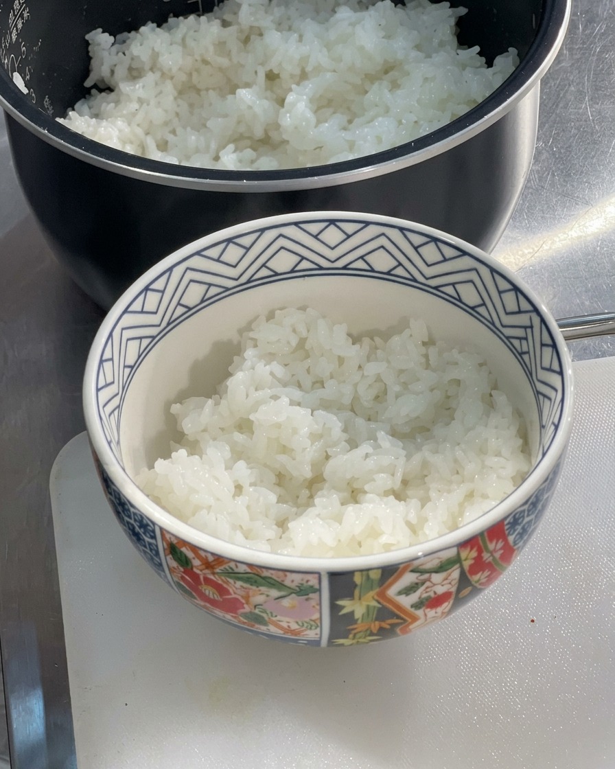 밥솥 옆 도마 위에 놓인 아기자기한 도자기 그릇에 갓 지은 촉촉하고 하얀 쌀밥이 반쯤 담겨 있는 모습.