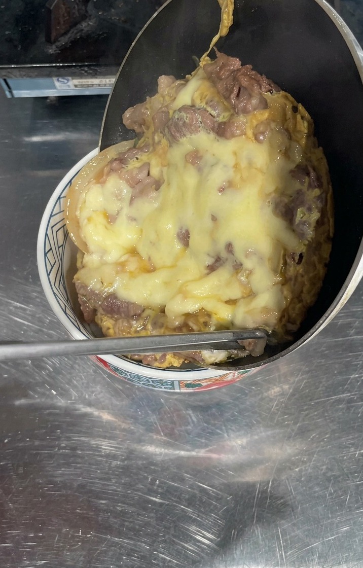 흰밥이 담긴 그릇 위로 프라이팬을 기울여 잘 졸여진 소고기와 황금빛으로 녹은 치즈 토핑을 그대로 미끄러뜨려 얹는 모습.
