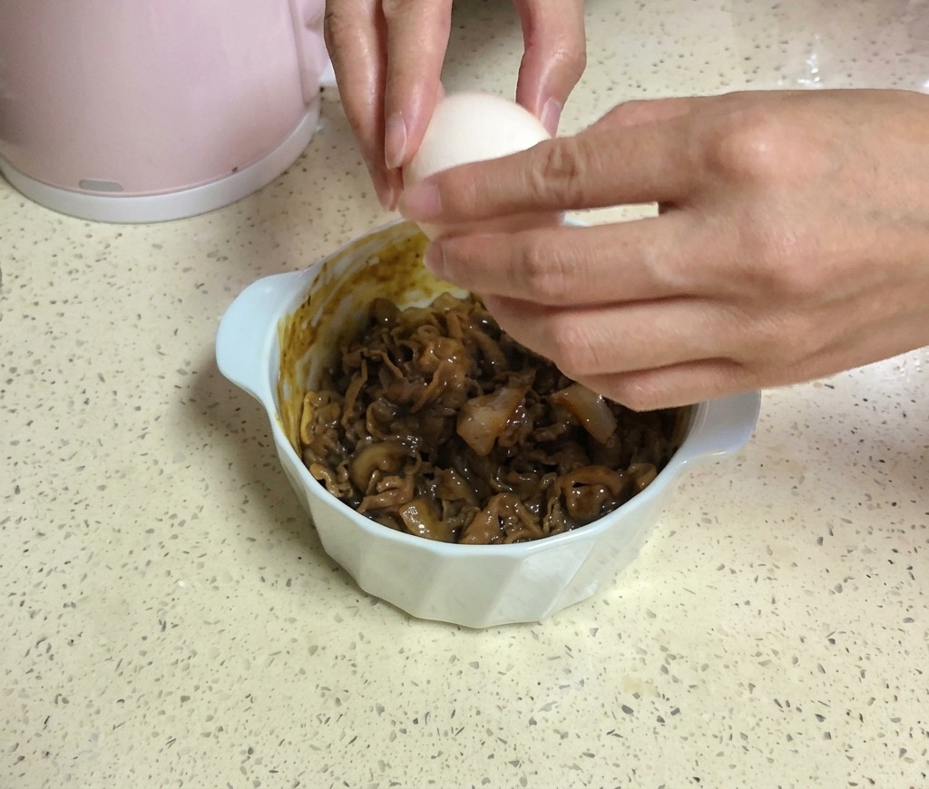 노란 치즈 두 장이 올라간 소고기 그릇 위로 양손을 이용해 신선한 날계란을 깨뜨려 넣는 모습.