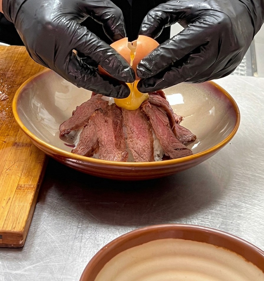 장갑을 낀 두 손으로 소고기 화산 꼭대기에 부드러운 온센타마고를 조심스럽게 깨뜨려 올리는 모습