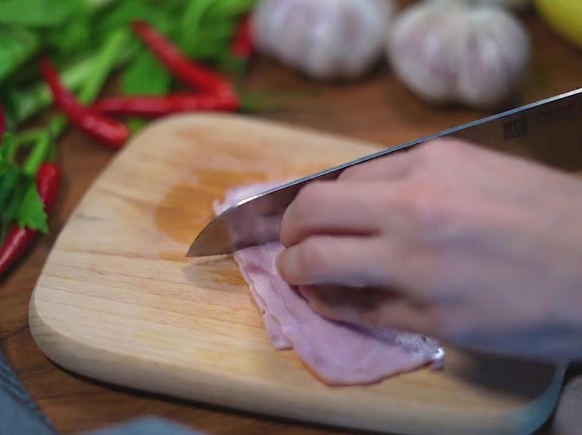 밝은 색 나무 도마 위에서 스테인리스 칼로 분홍빛 생베이컨을 한 입 크기의 정사각형으로 써는 모습.
