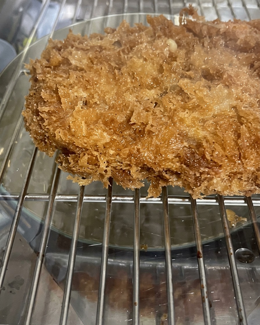 식힘망 위에서 기름을 빼며 휴지 중인 바삭하고 거친 질감의 빵가루가 돋보이는 돈가스 확대 모습.