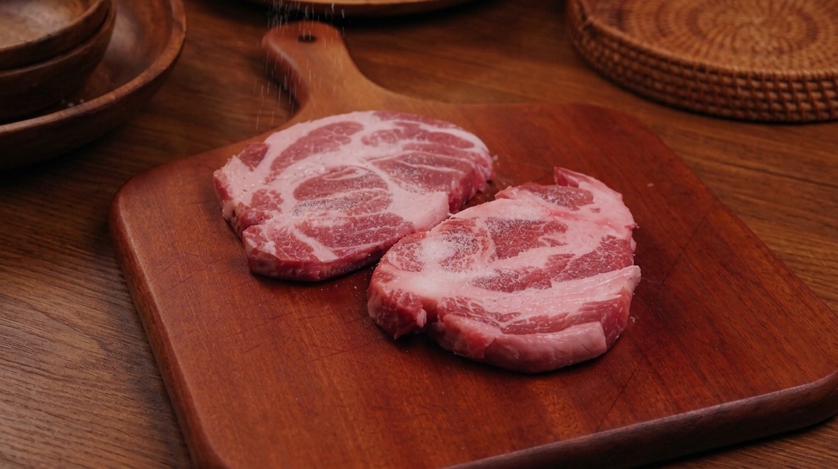 나무 도마 위에 올려진 두 조각의 돼지고기 위로 소금과 후추를 뿌리는 모습.