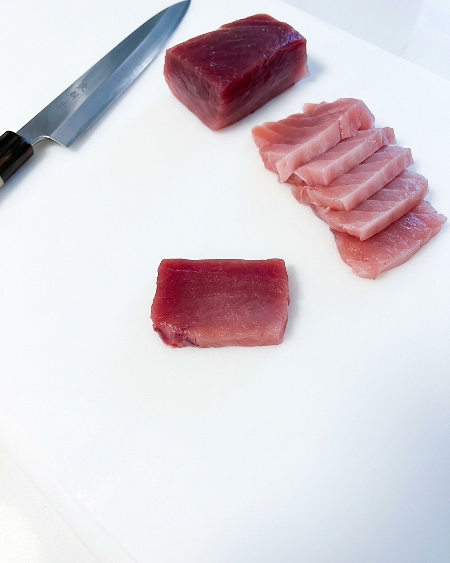 하얀 도마 위에 참치 덩어리와 일본식 식칼이 놓여 있고, 그 옆에 직사각형으로 썰린 분홍빛 참치 뱃살이 가지런히 놓인 모습.