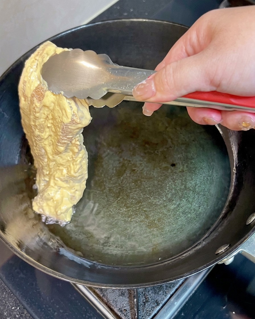 반죽 옷을 입힌 삼겹살 한 조각을 금속 집게를 이용해 팬 안의 뜨거운 기름 속으로 조심스럽게 넣는 모습.