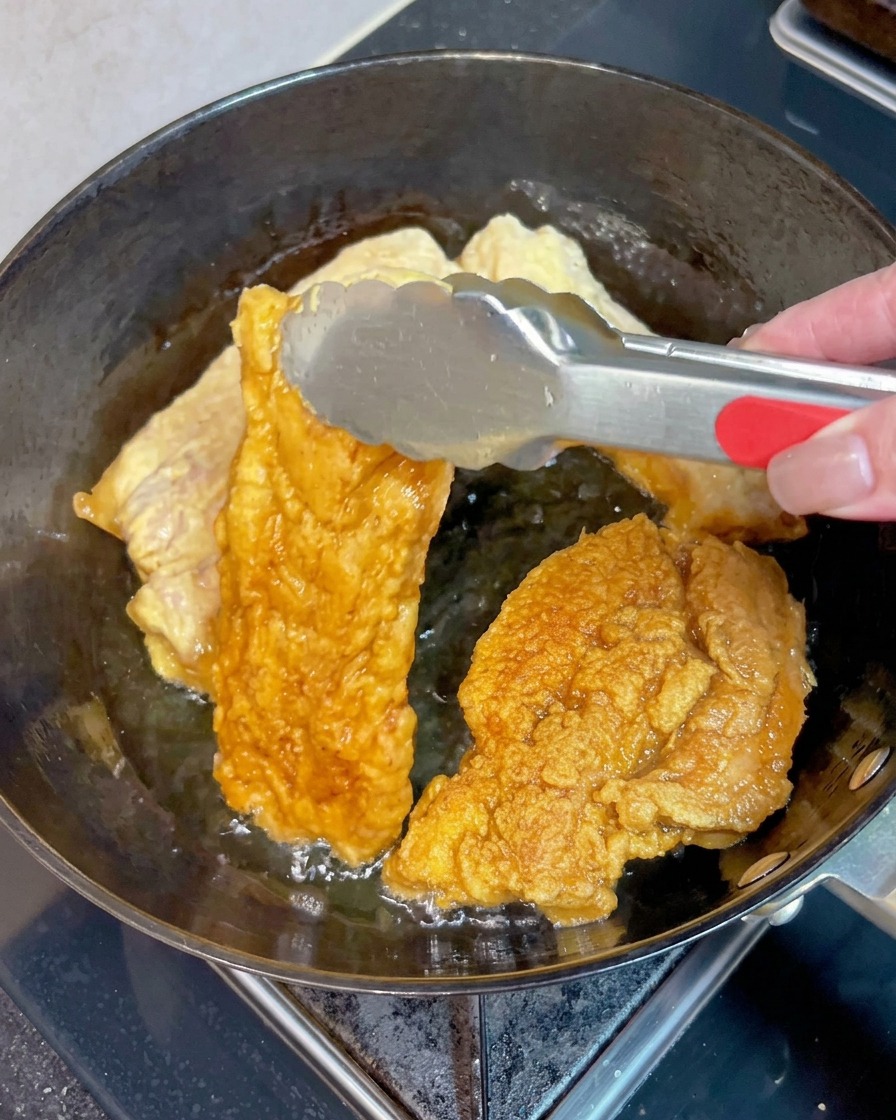 지글지글 끓는 뜨거운 기름 속에서 금속 집게로 노릇노릇하고 바삭하게 튀겨진 삼겹살 조각을 뒤집는 모습.