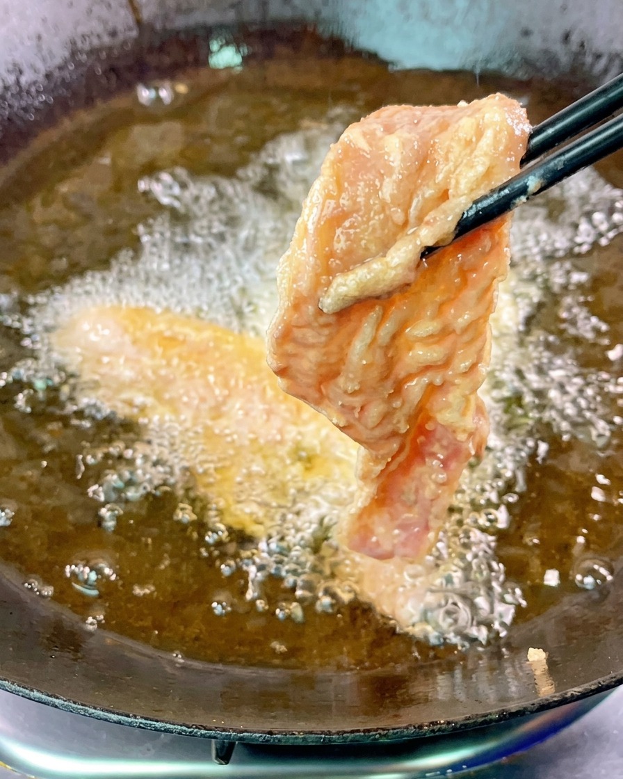 검은색 젓가락으로 반죽 옷을 입힌 삼겹살 한 조각을 자글자글 끓는 뜨거운 기름에 넣는 모습.