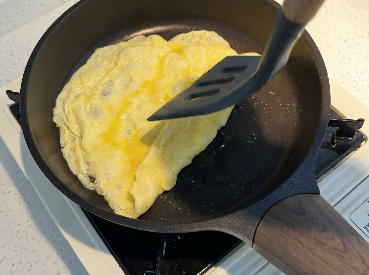 달궈진 코팅 팬에서 회색 뒤집개로 얇게 익은 노란 달걀판을 조심스럽게 접고 있는 모습.