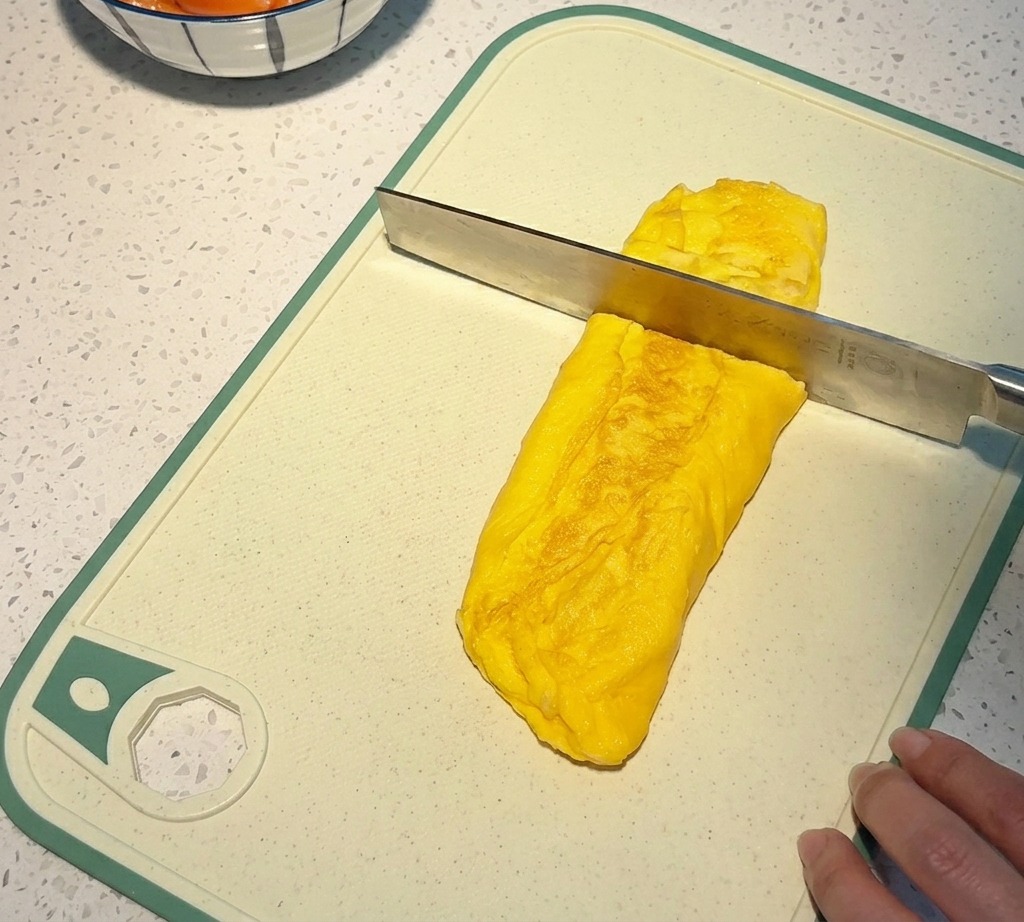 흰색 도마 위에서 두툼하고 노란 달걀말이를 칼로 썰어 작은 정사각형 큐브 모양으로 만드는 모습.