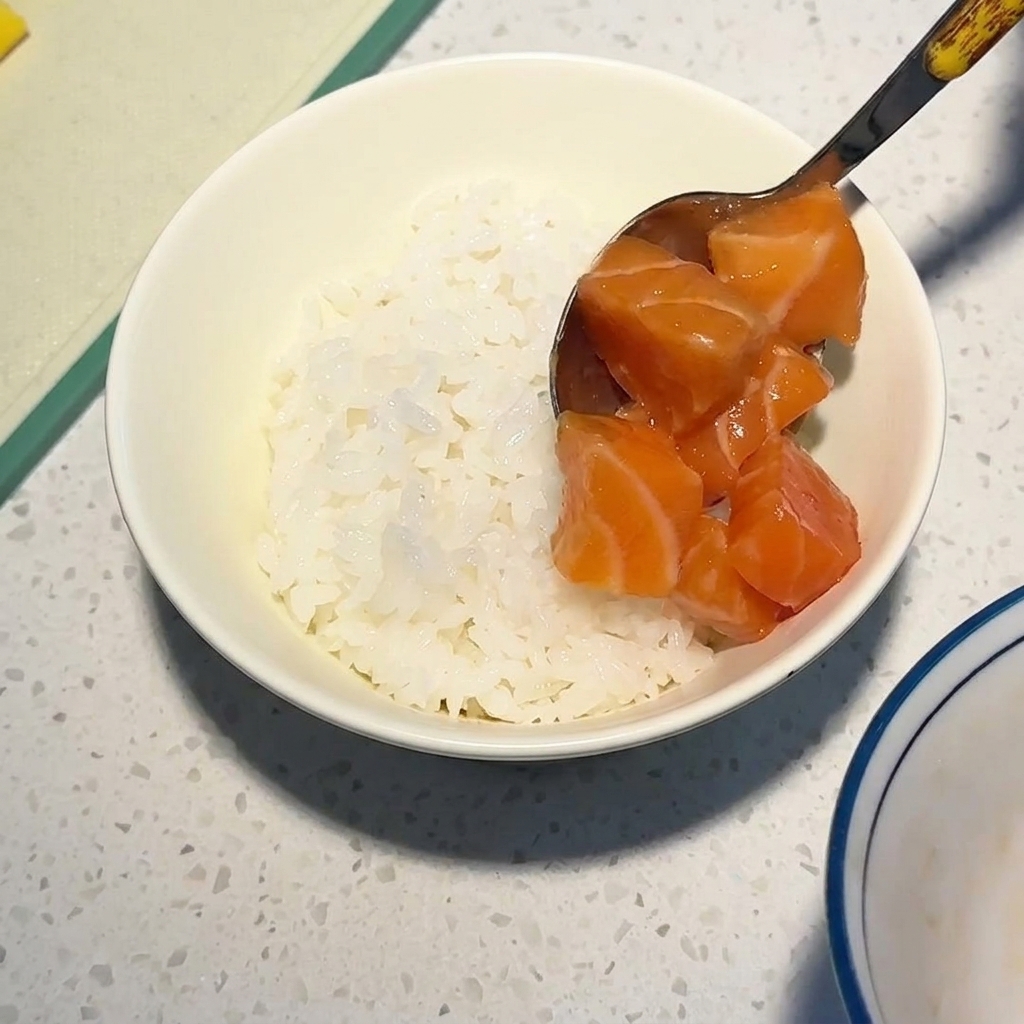 쇠숟가락으로 윤기가 흐르는 생연어 큐브를 볼에 담긴 흰밥 위에 올리는 모습.
