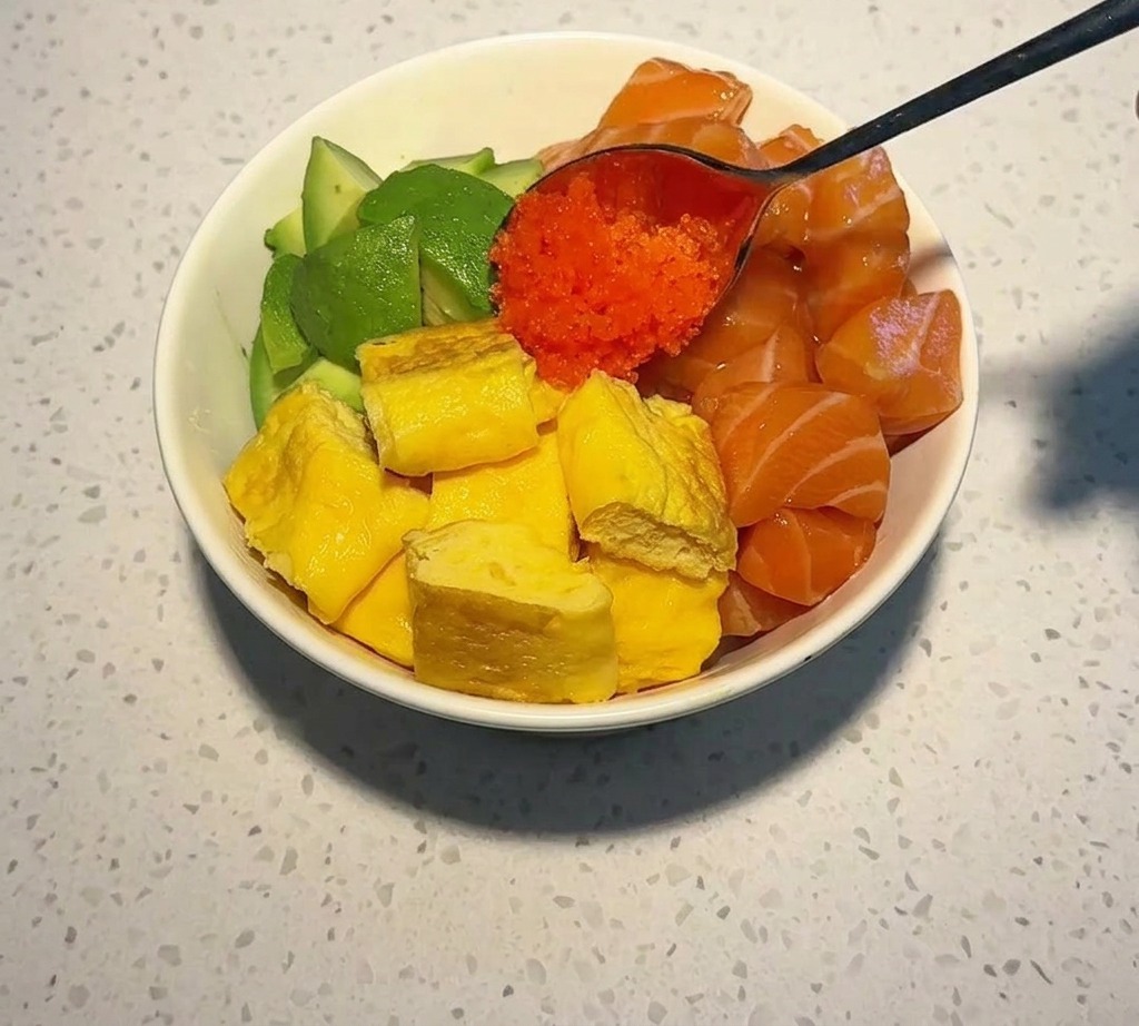 연어, 아보카도, 달걀말이가 채워진 그릇 중앙에 쇠숟가락으로 선명한 주황색 날치알을 올리는 모습.