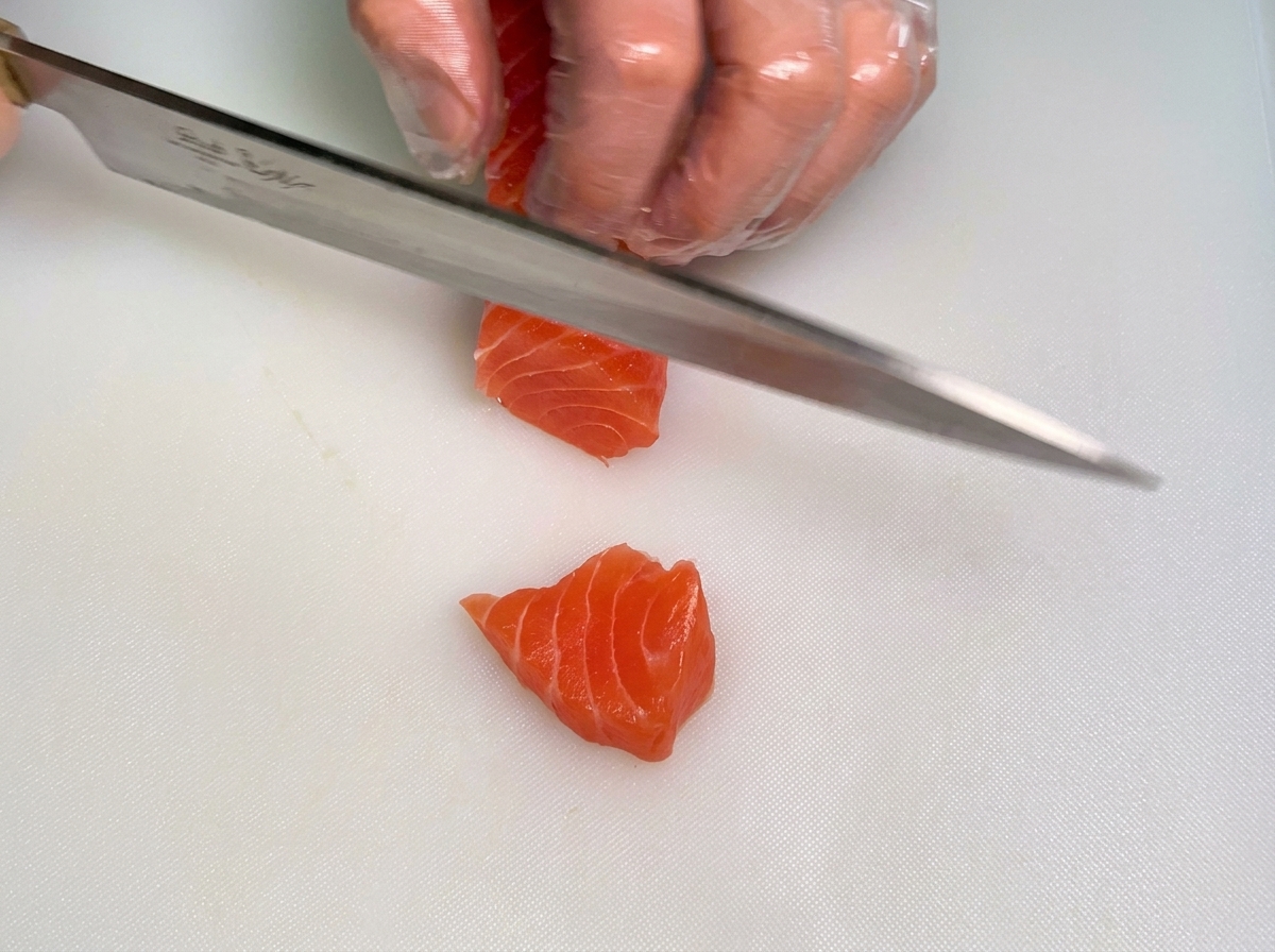 흰색 도마 위에서 한 손으로는 생연어 덩어리를 잡고 다른 손으로는 칼을 이용해 두툼한 막대 모양으로 썰어내는 셰프의 장갑 낀 손