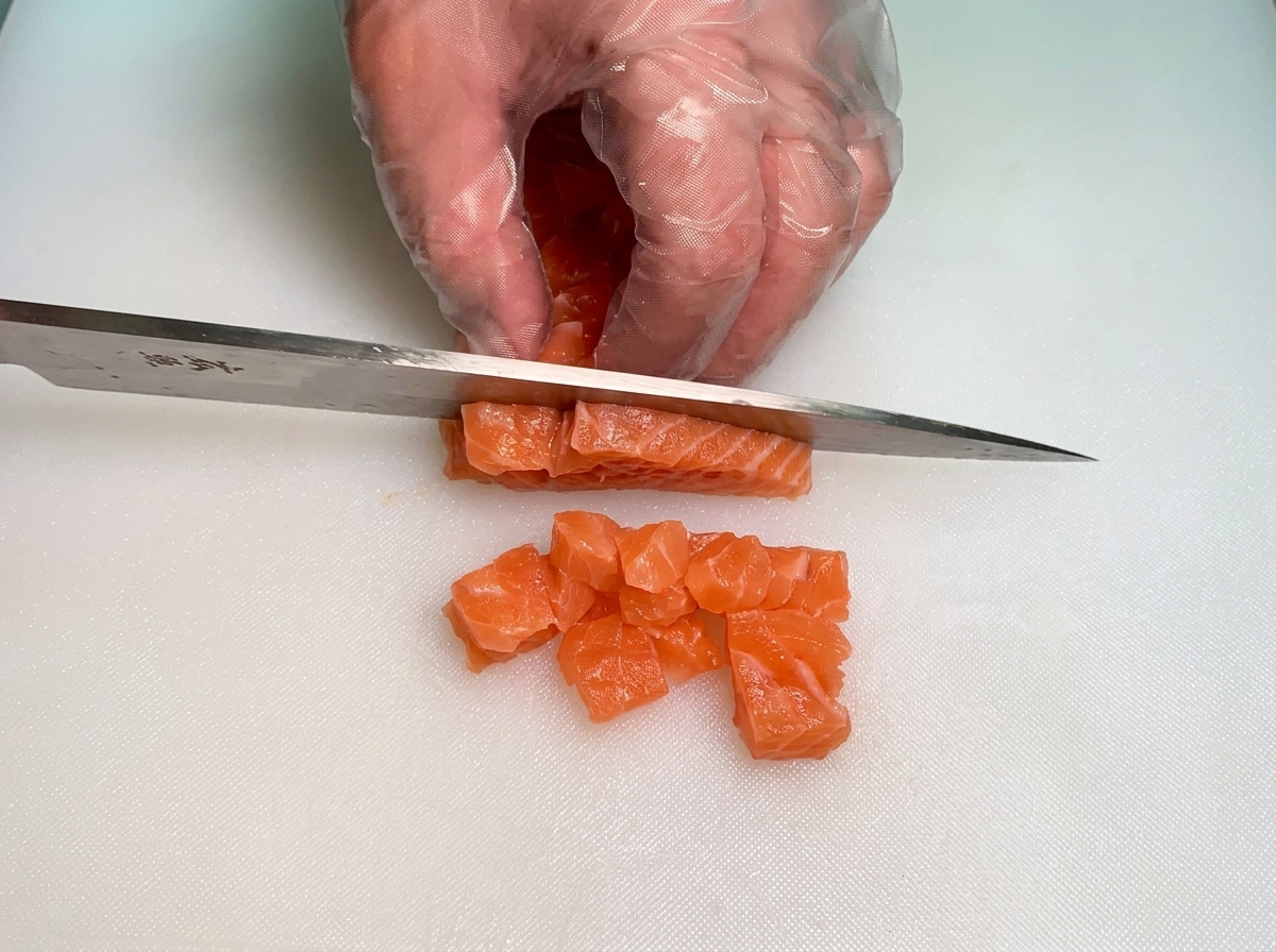 장갑을 낀 손으로 칼을 사용해 선명한 주황색 생연어 막대를 일정한 크기의 작은 큐브 모양으로 깍둑썰기하는 모습