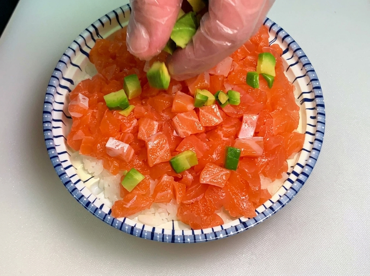 깍둑썰기한 연어와 흰밥이 깔린 대접 위에 투명 장갑을 낀 손으로 초록색 아보카도 큐브를 골고루 뿌려주는 모습