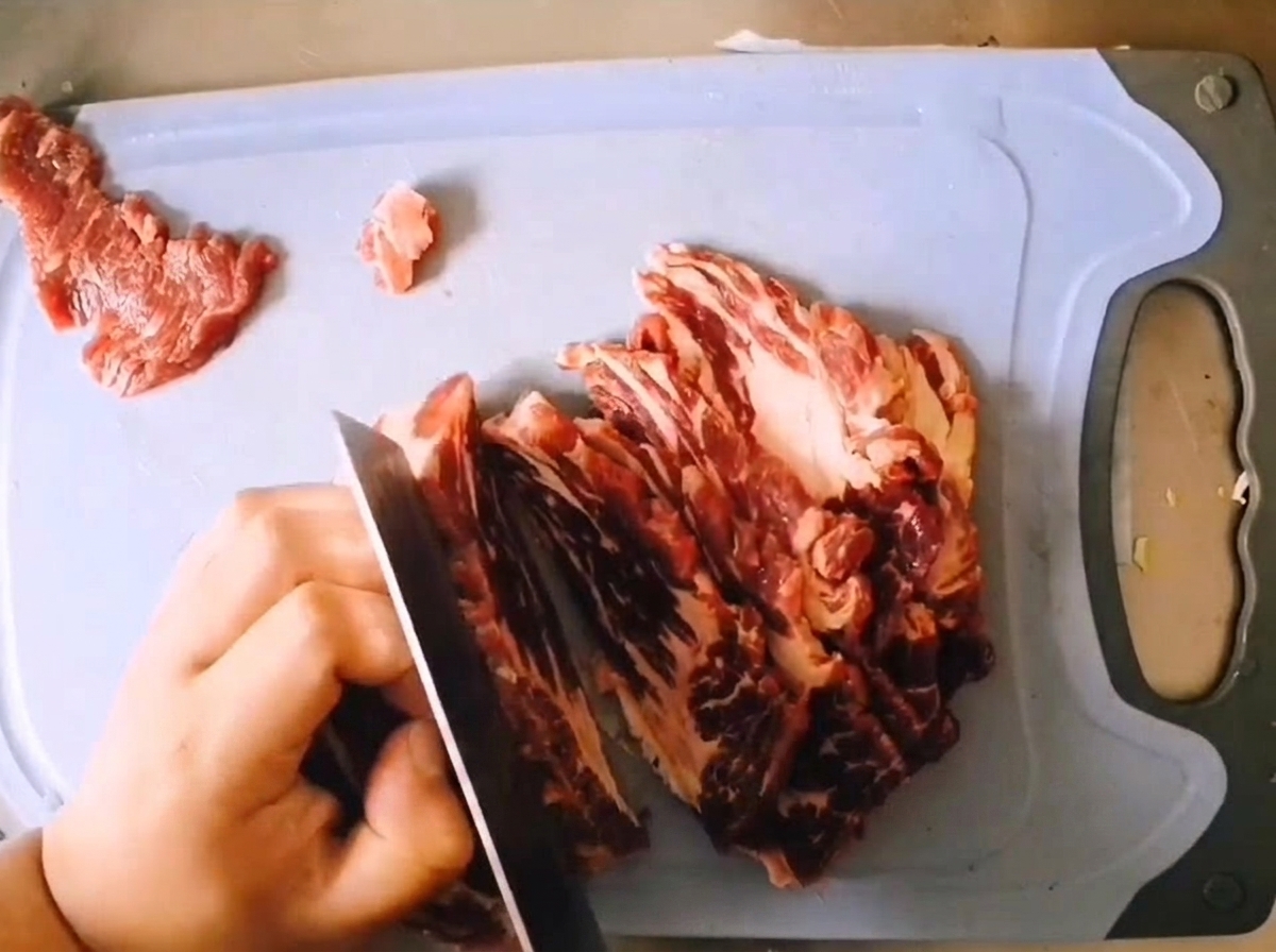 연한 푸른색 도마 위에서 날카로운 칼로 마블링이 있는 생 소고기 목심을 두툼하게 썰어내는 모습.