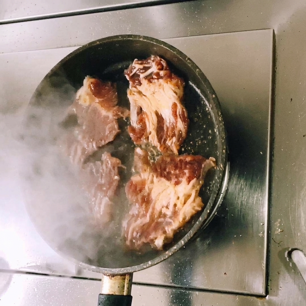 달구어진 평평한 팬 위에서 양념된 소고기가 구워지며 김이 피어오르고 고기 겉면이 갈색으로 변해가는 모습.