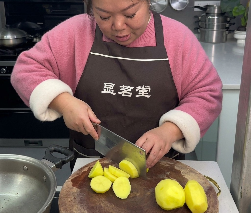갈색 앞치마를 두른 여성이 둥근 나무 도마 위에서 큰 주방 칼로 껍질을 깐 노란 감자를 돌려깎기 스타일로 썰고 있는 모습.