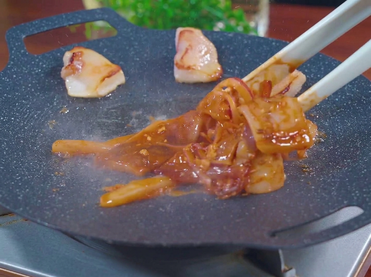 연기가 나는 뜨거운 검은색 그릴 팬 위에 녹은 돼지 비계 조각들과 함께 양념된 오징어와 양파 슬라이스를 집게로 올리는 모습.