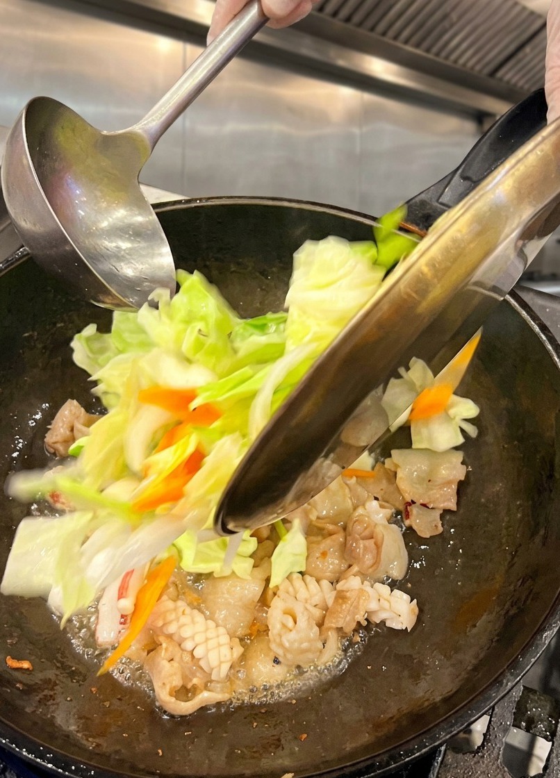 삼겹살과 오징어가 익어가고 있는 뜨거운 웍에 금속 접시에 담긴 먹기 좋게 썬 양배추와 당근을 넣는 모습.