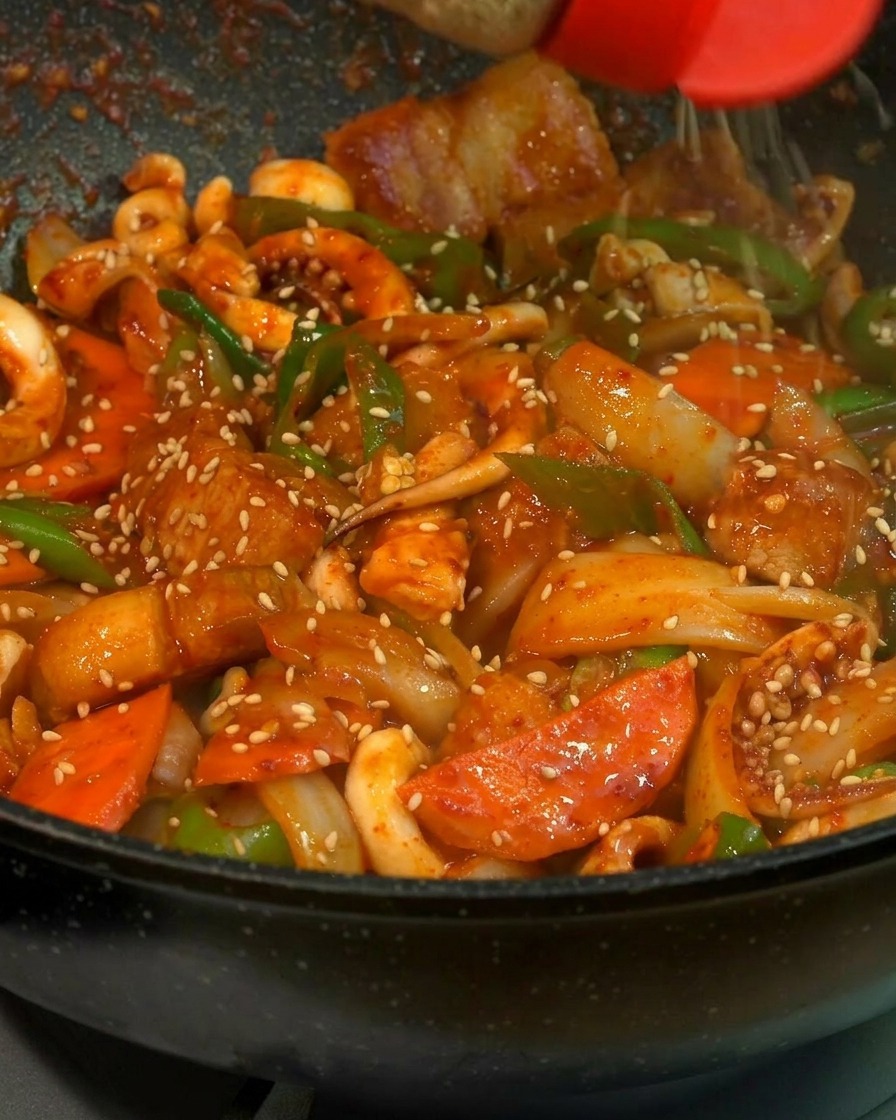 검은색 팬에 담긴 먹음직스러운 오삼불고기 위에 빨간 뚜껑의 양념통에서 흰 참깨를 솔솔 뿌리는 근접 샷