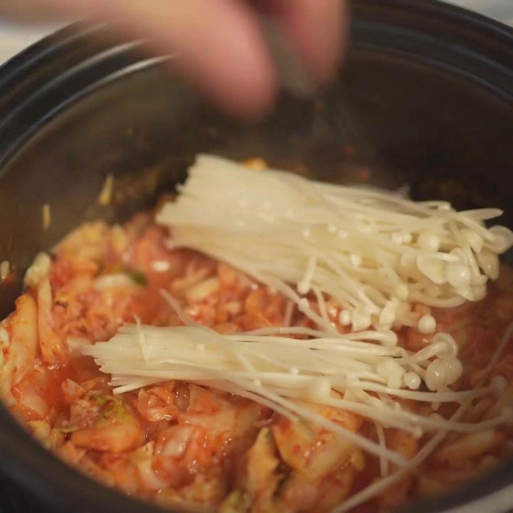 뚝배기 속 볶은 김치 위에 밑동을 자른 신선한 팽이버섯 한 묶음을 가지런히 올리는 모습
