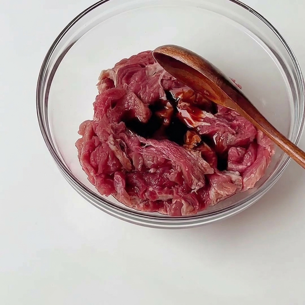 유리 볼에 담긴 얇게 썬 소고기에 간장 양념을 넣고 나무 숟가락으로 고르게 버무리는 모습.