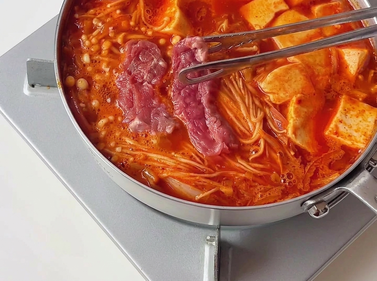 두부와 버섯 옆으로 붉게 끓어오르는 찌개 속에 밑간한 핑크빛 소고기를 집게로 펼쳐 넣는 모습.