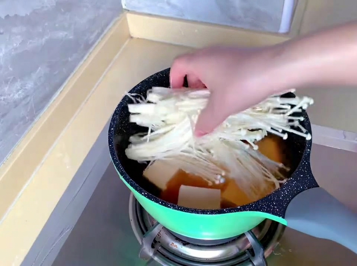 양념 국물과 순두부가 담긴 냄비에 하얗고 긴 팽이버섯 한 묶음을 손으로 넣고 있는 모습