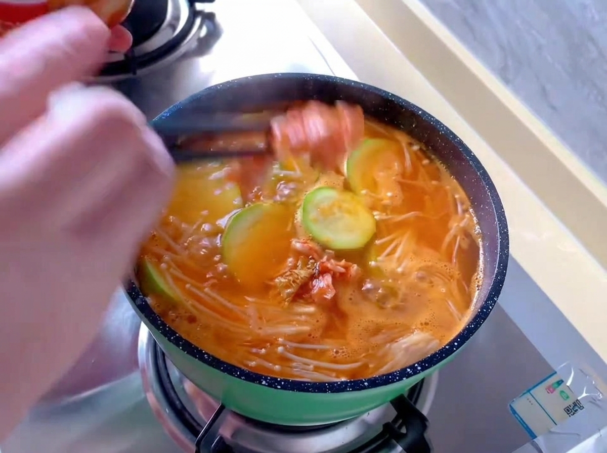 팽이버섯과 애호박이 끓고 있는 주황색 김치찌개 냄비에 젓가락으로 빨간 배추김치를 넣는 모습
