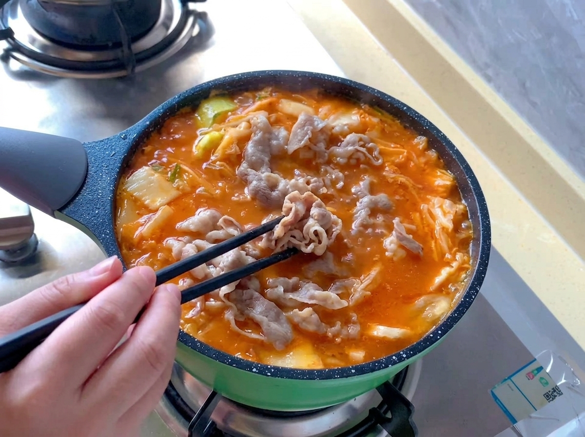 보글보글 끓어오르는 빨간 김치찌개 국물 속에 데쳐둔 소고기 롤을 검은색 젓가락으로 펼쳐 넣는 모습