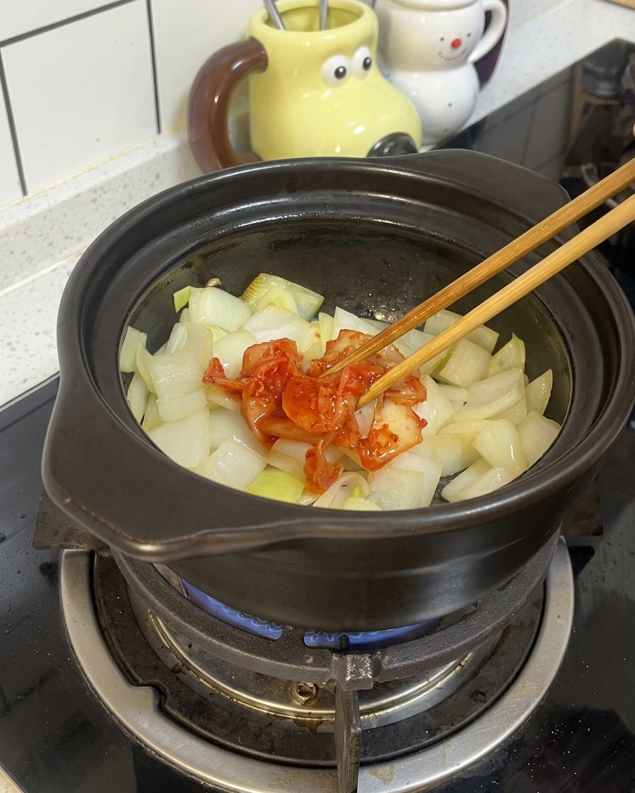 뚝배기에 볶은 양파 위로 붉은 신김치를 넣고 있는 모습.