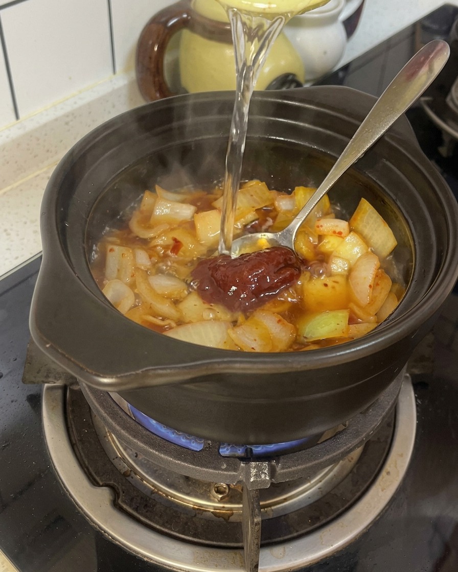 양념한 김치와 양파가 담긴 뚝배기에 물을 붓고 있는 모습.