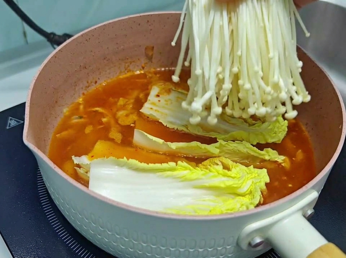 진한 주황빛 김치찌개 국물에 깨끗하게 손질된 흰 팽이버섯 한 묶음을 넣는 모습