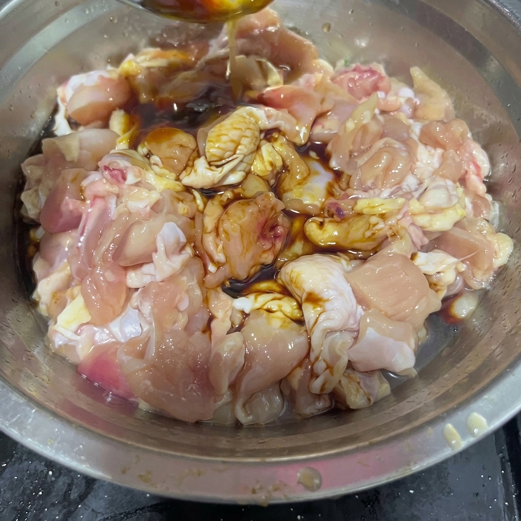 생 닭고기 조각과 진한 소스들이 담긴 볼에 스테인리스 스푼으로 맛술을 붓고 있는 모습.