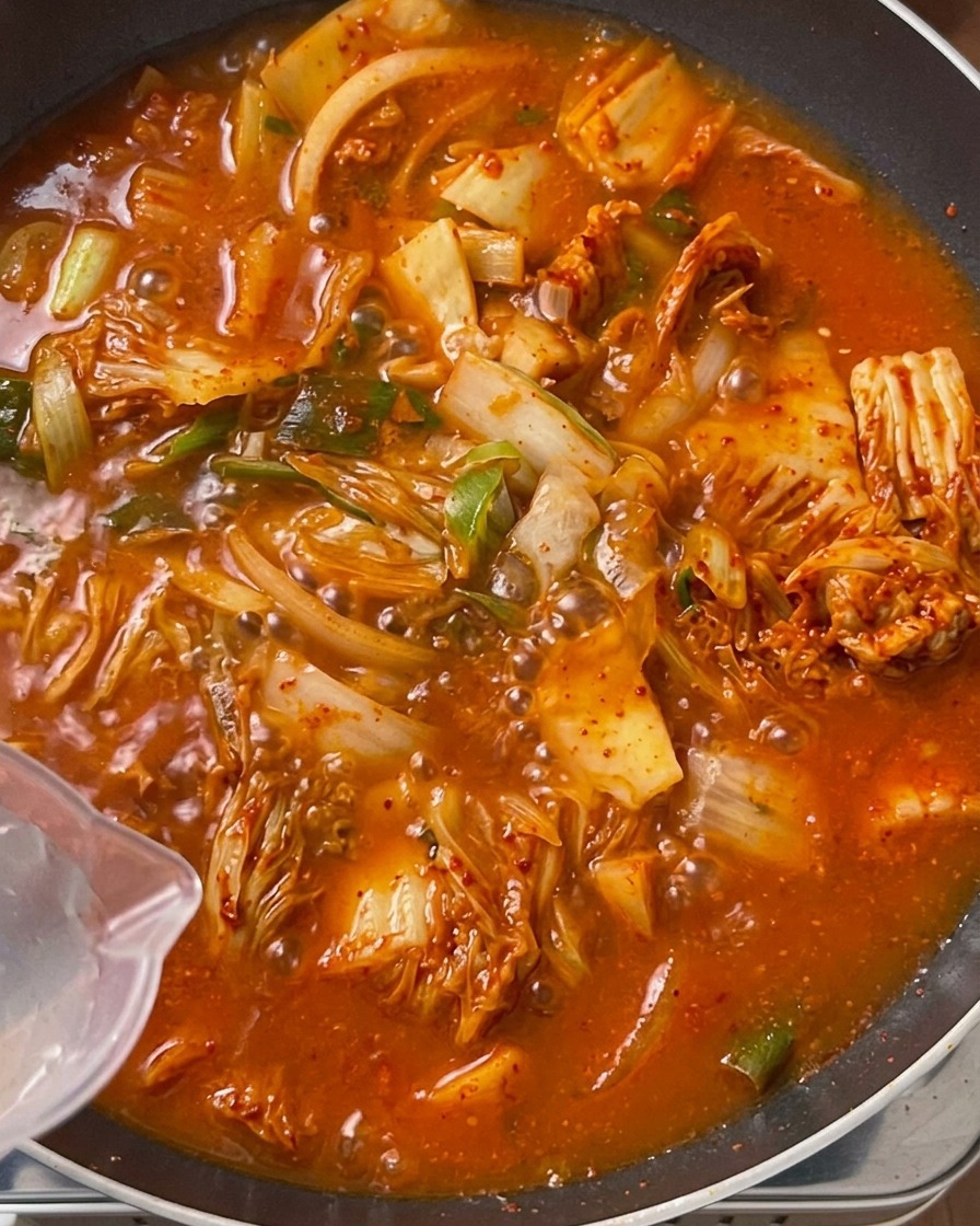 보글보글 끓어오르는 주황빛 김치 고기 볶음이 가득 찬 큰 팬에 투명한 계량컵으로 맑은 물을 붓는 모습