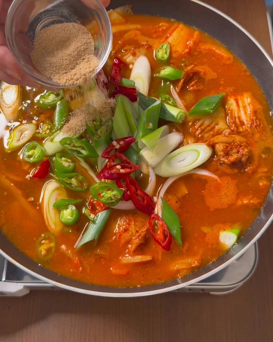 고추와 대파가 가득한 끓는 찌개 표면 위로 작은 투명 종지에 담긴 갈색 쇠고기 조미료 가루를 뿌리는 모습