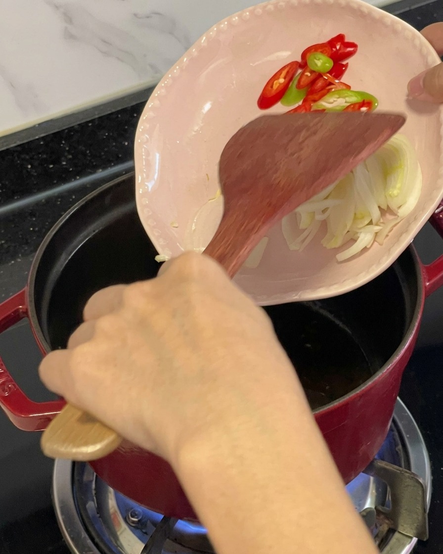오일을 두른 빨간 냄비에 나무 주걱을 사용하여 분홍색 작은 접시에서 썬 홍고추, 청고추, 향신 채소를 넣고 볶는 모습.