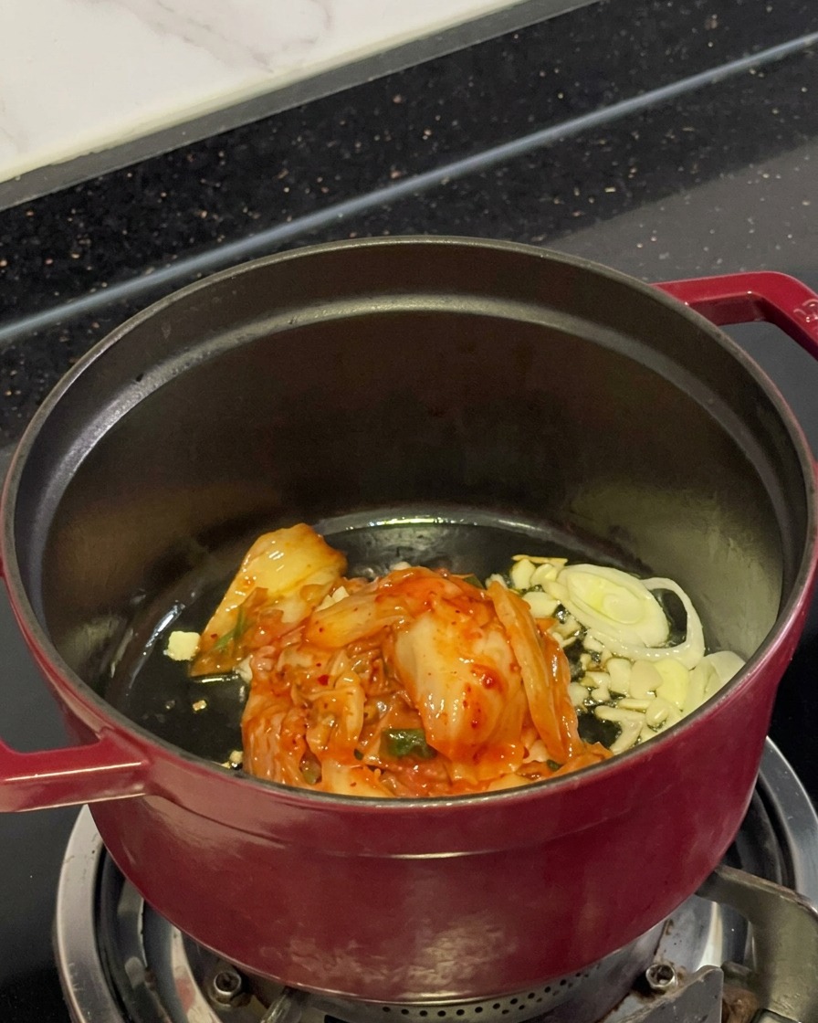 냄비 안에서 오일에 볶아지고 있는 먹기 좋게 자른 빨간 김치와 다진 마늘, 대파의 모습.