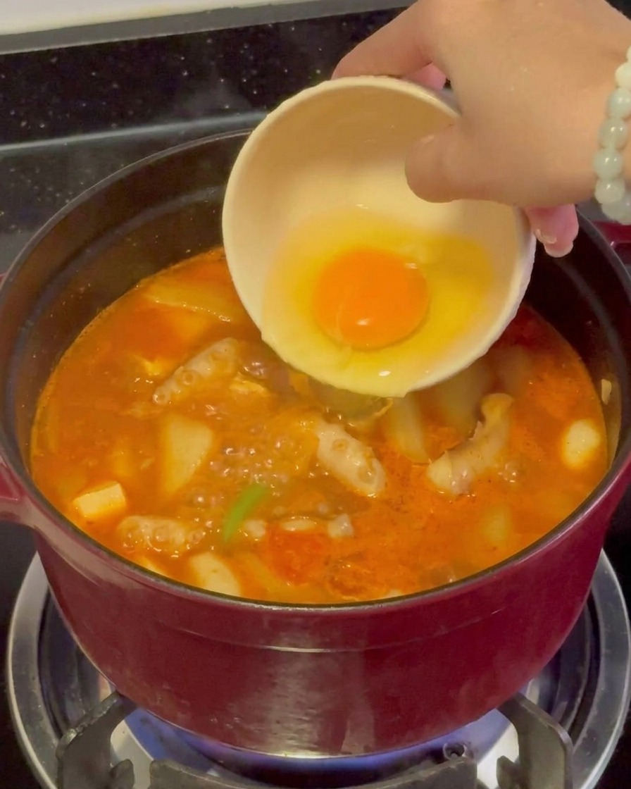 강하게 끓고 있는 빨간 해물 찌개 중심에 선명한 노란 노른자가 살아있는 날달걀을 조심스럽게 붓는 모습.