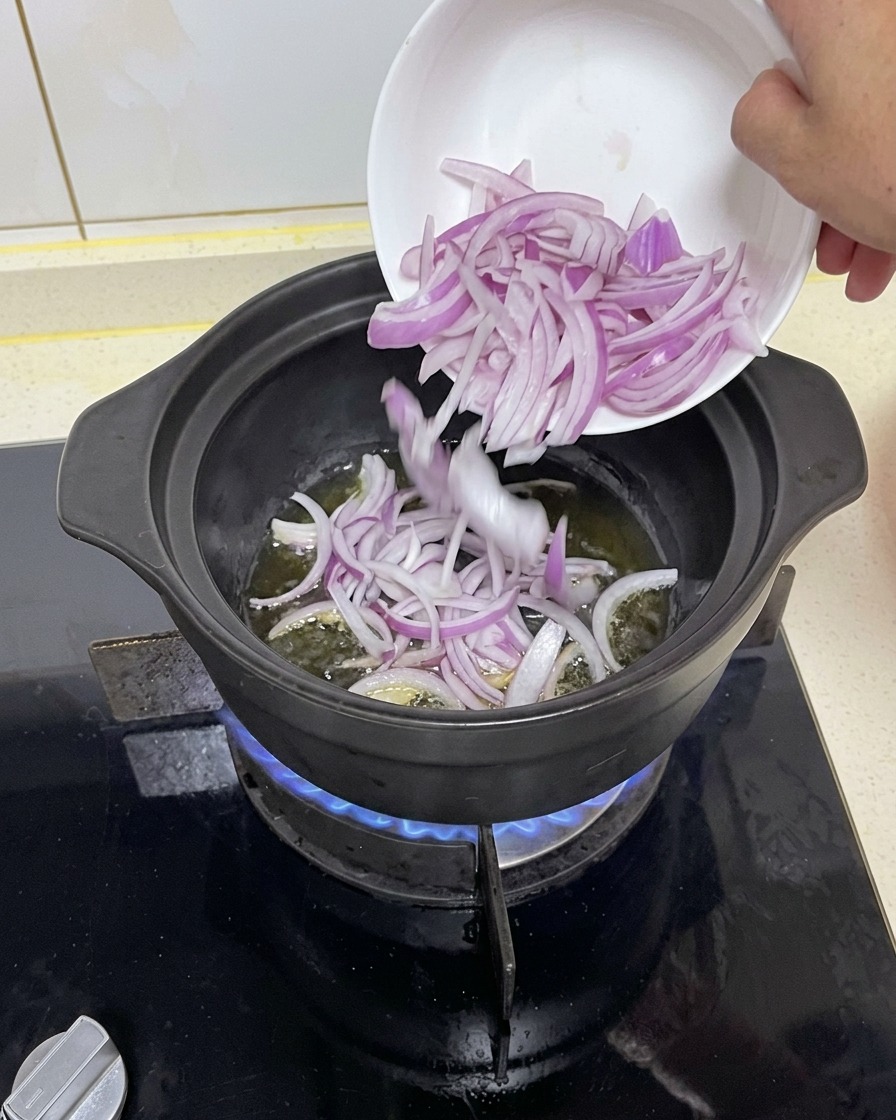 파란 가스불 위 달궈진 검은 냄비에 하얀 작은 접시로부터 채 썬 적양파를 넣고 기름에 볶는 모습.
