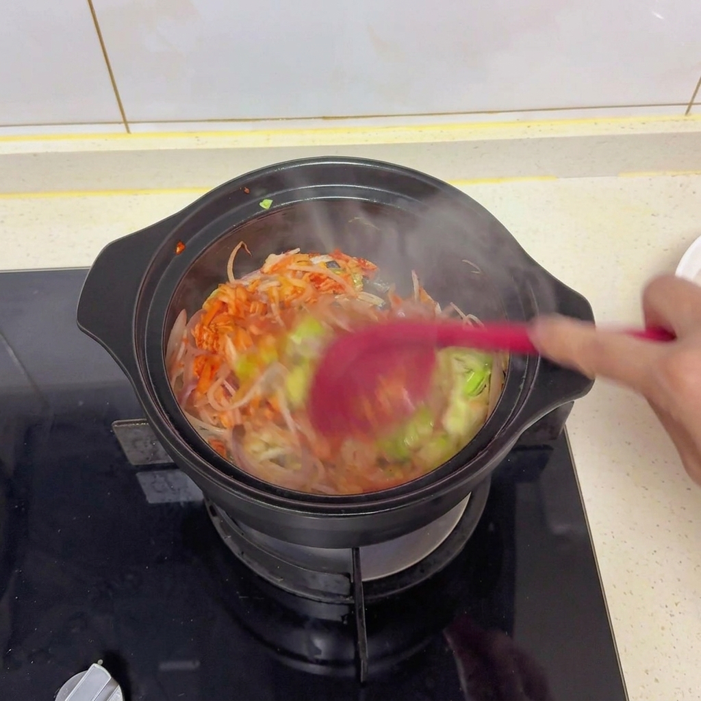 빨간 주걱으로 검은 냄비 속 채소와 김치를 볶으며 소금และ 간장 등의 양념을 골고루 섞는 모습.