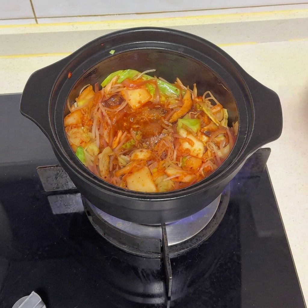 볶아진 채소와 김치 위에 다양한 양념과 붉은 고춧가루가 올려져 있는 검은 냄비의 모습.