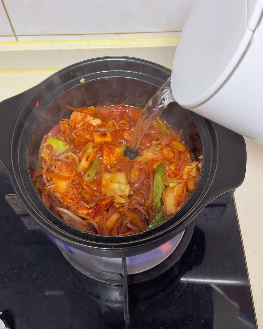 가스레인지 위 검은 뚝배기에 흰 전기포트로 뜨거운 물을 부어 볶은 재료들과 섞이며 빨간 국물이 만들어지는 모습.