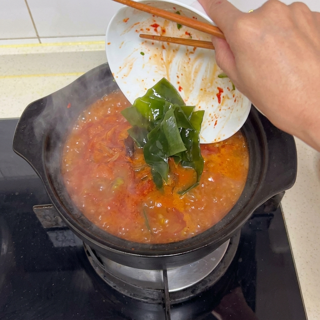 보글보글 끓어오르는 새빨간 찌개 국물에 젓가락을 이용해 흰 접시에 담긴 짙은 초록색 다시마 조각들을 넣는 모습.